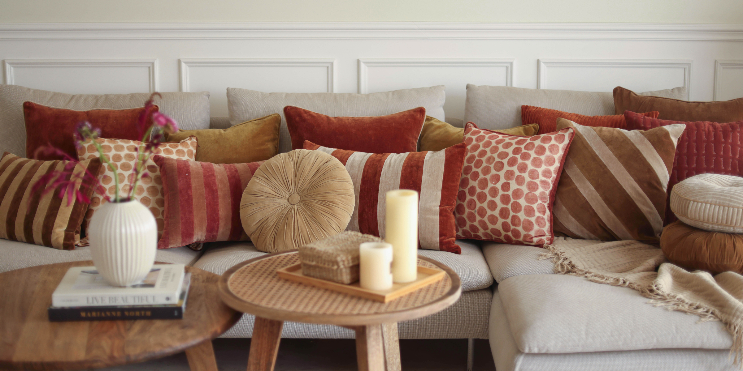 A stylish living room decoration featuring a beige sofa styled with an assortment of fall themed throw pillows, including square couch pillows and velvet sofa pillow covers in warm rust, mustard, and beige tones.